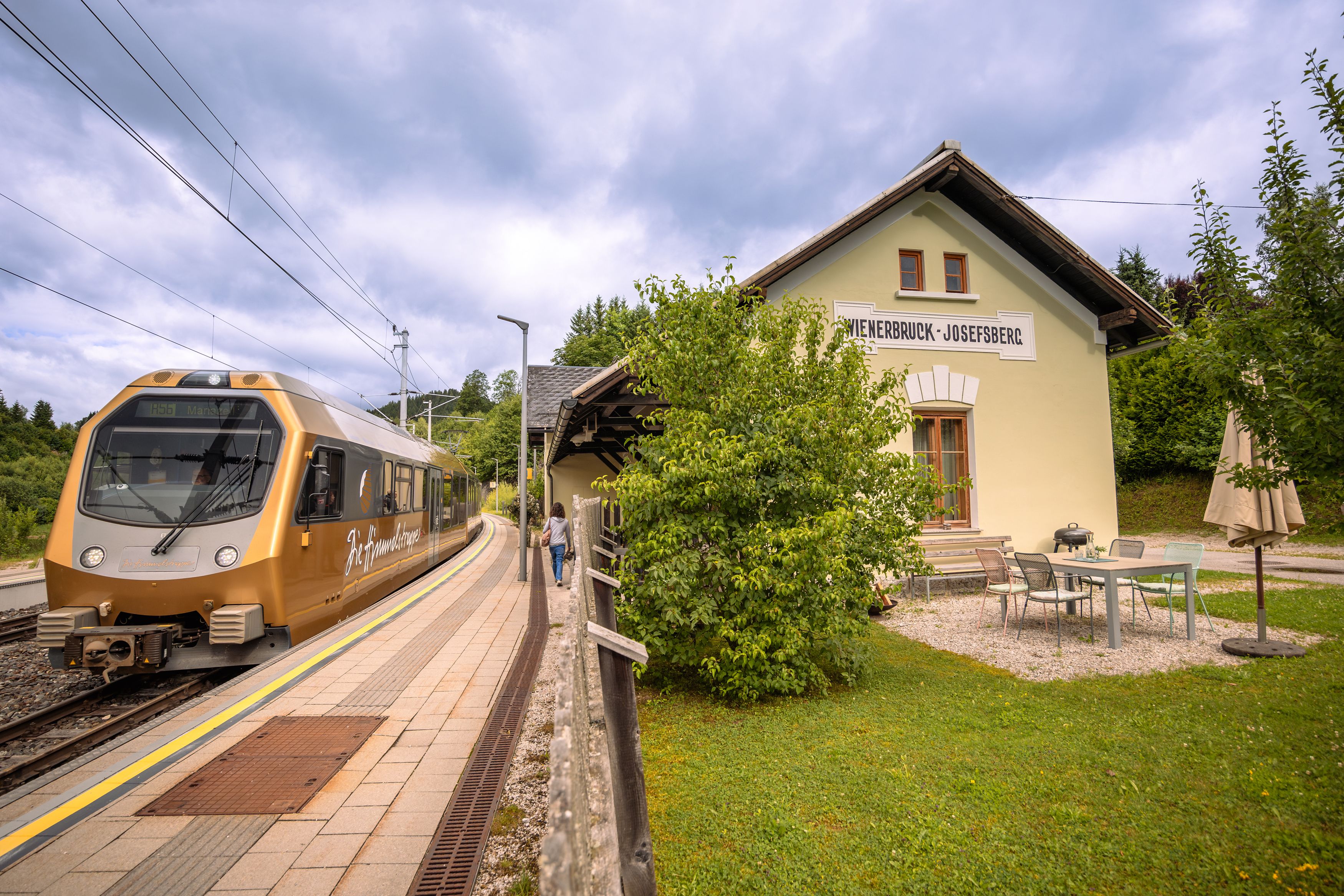 A golden train stops at Wienerbruck-Josefsberg station, surrounded by green countryside and a small outdoor seating area.