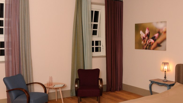 A cozy room with two armchairs, a small table, a bed and a lamp. A picture with a flower motif hangs on the wall.
