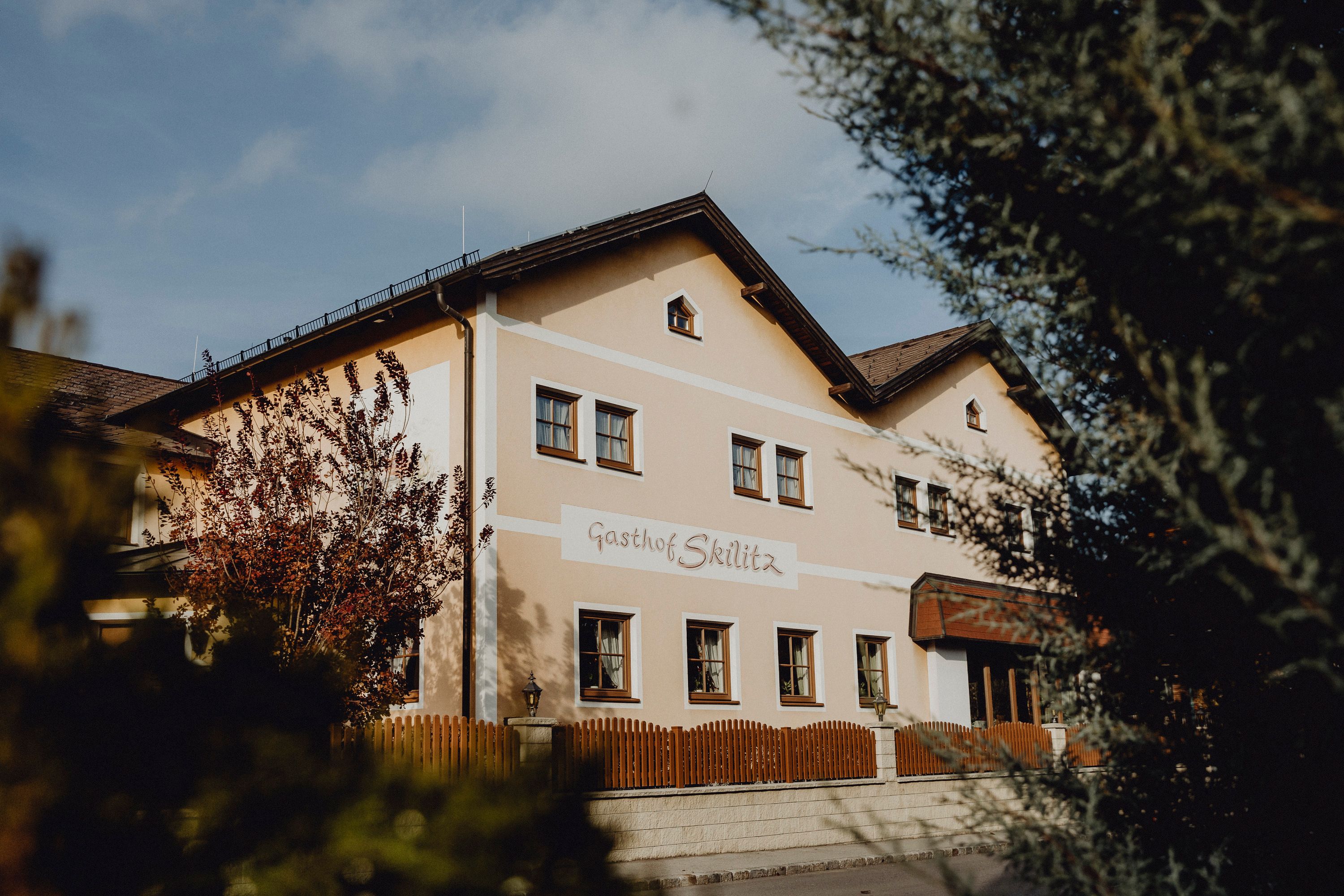 A traditional inn with the inscription 'Gasthof Skilitz', surrounded by trees and a wooden fence.