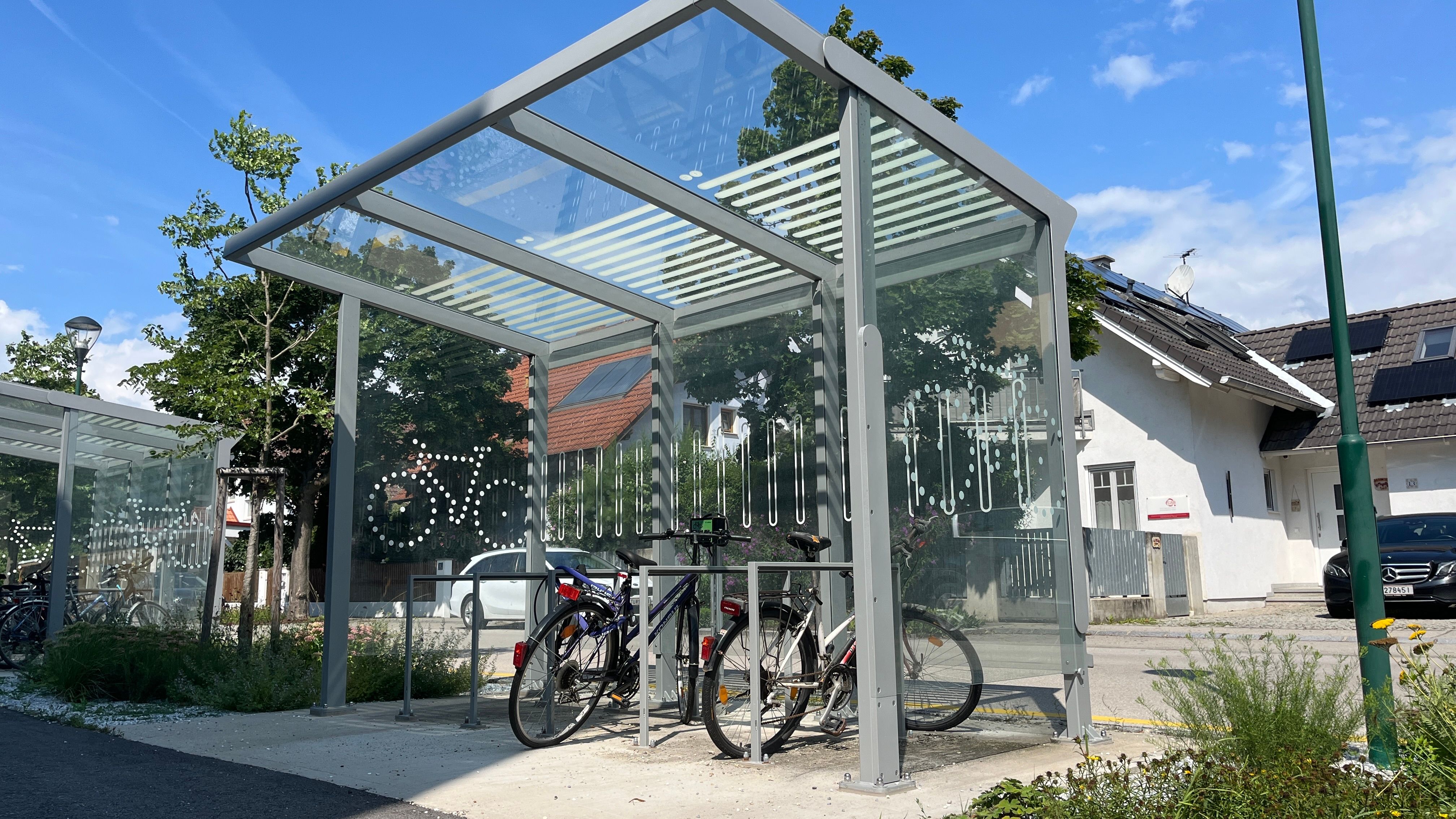 E-bike charging station with covered area and bicycles, in the background a house with solar panels.