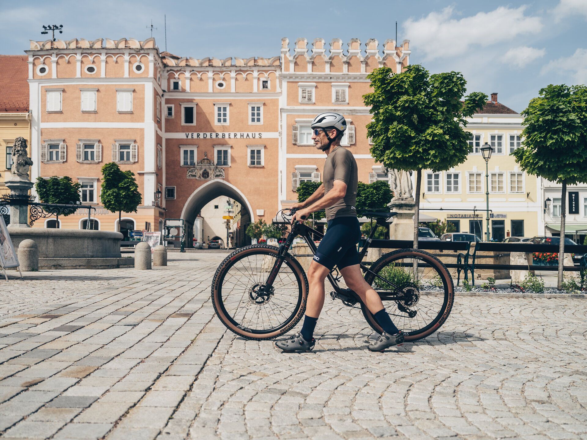 Ein Radfahrer genießt die malerische Atmosphäre der Stadt, während er sein Fahrrad durch die charmanten Gassen schiebt. Die historischen Gebäude und die sanften Hügel im Hintergrund laden zu einer Erkundungstour ein, die die Schönheit der Region Weinviertel offenbart.