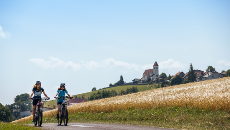 Two people riding e-bikes on a rural road, a village with a church in the background.
