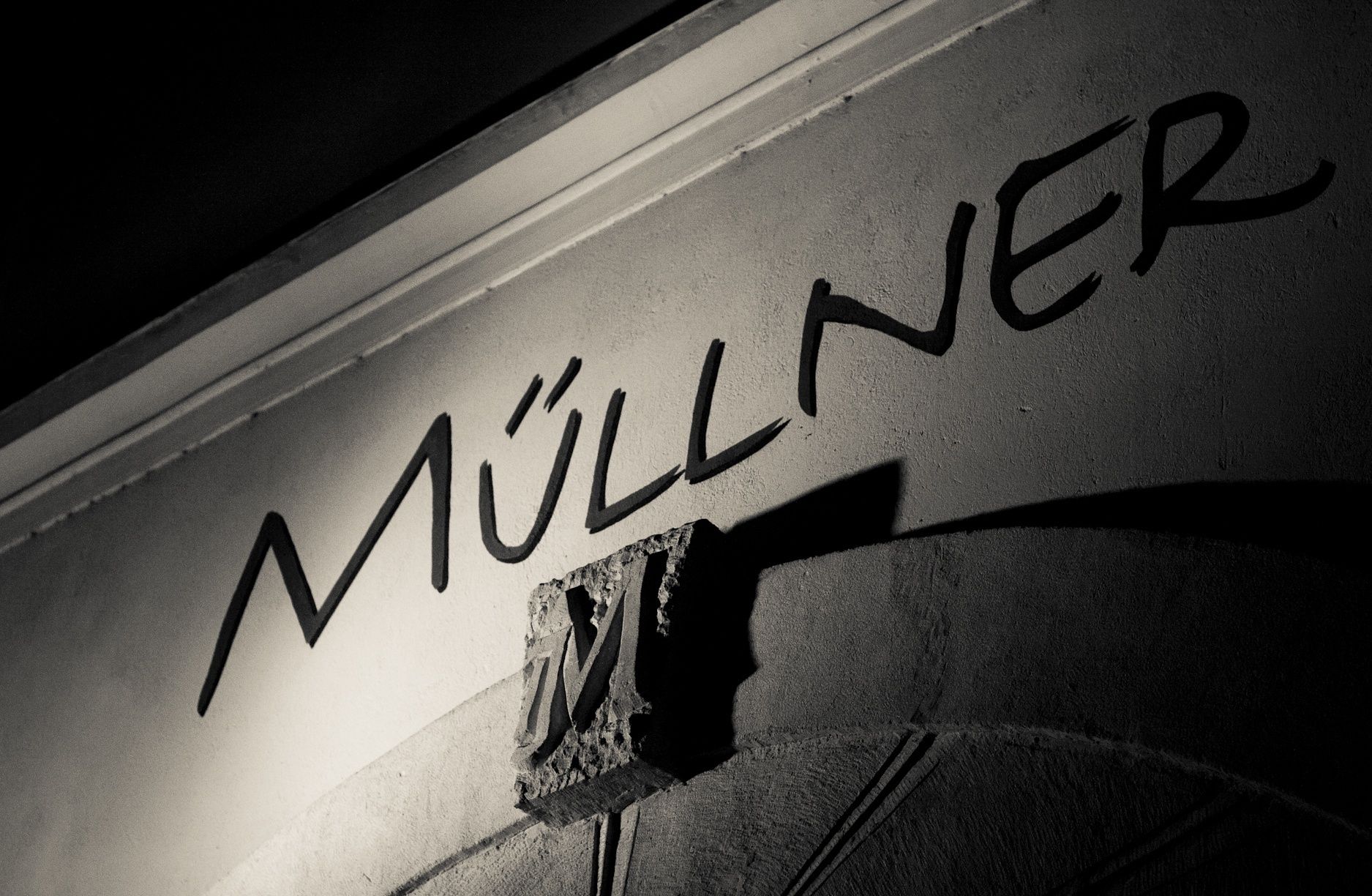 Black and white photo of a lettering 'Müllner' on a wall with a decorative 'M'.