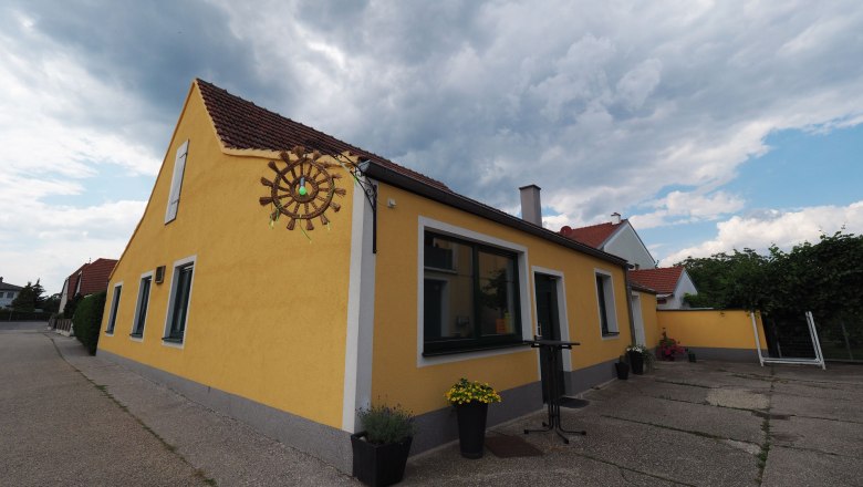Yellow building with red roof tiles and a decorative wheel on the wall, surrounded by plants and a cloudy sky.
