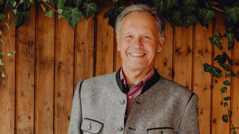 A smiling man in traditional dress in front of a wooden wall with ivy.