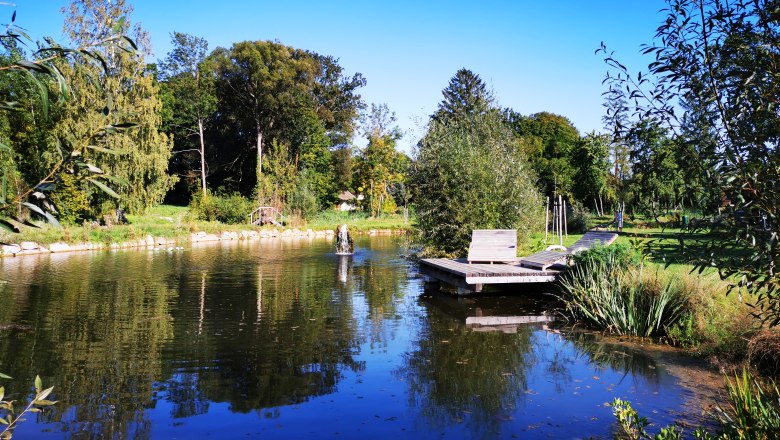 A tranquil pond with wooden loungers and lush vegetation under a blue sky.