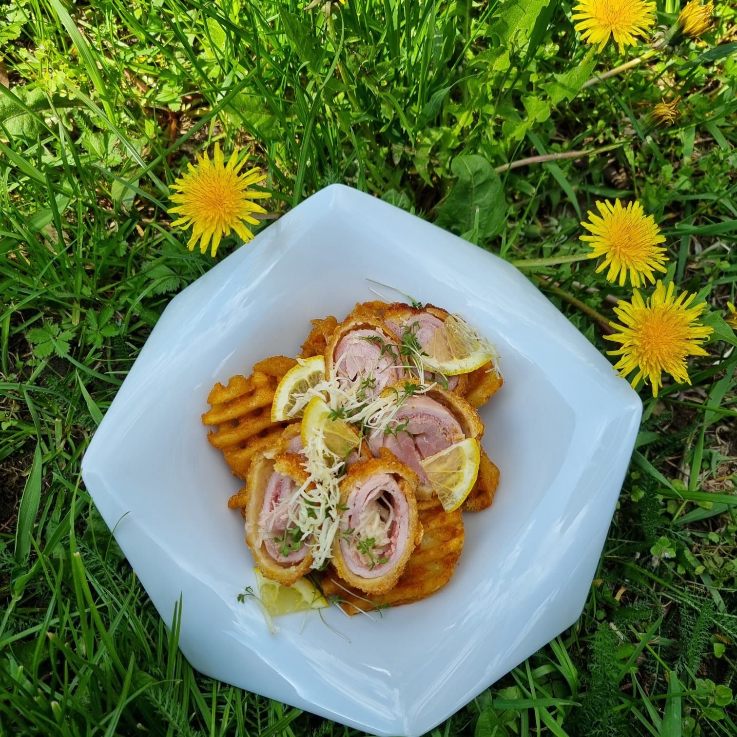 A plate of cordon rolls and waffle fries, garnished with lemon slices and cress, lies on a meadow of dandelions.