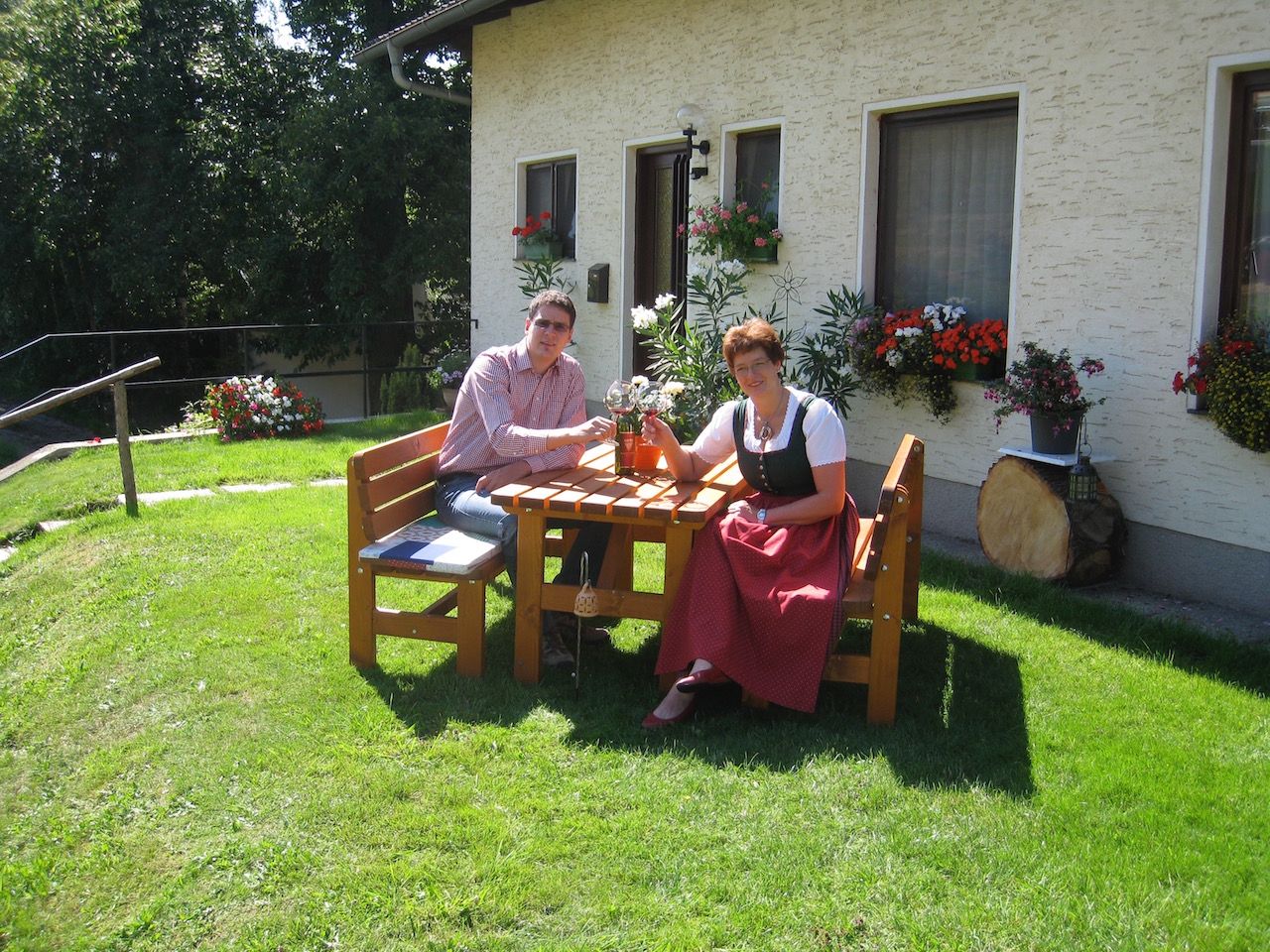 Two people are sitting at a wooden table in the garden in front of a house with flowers.