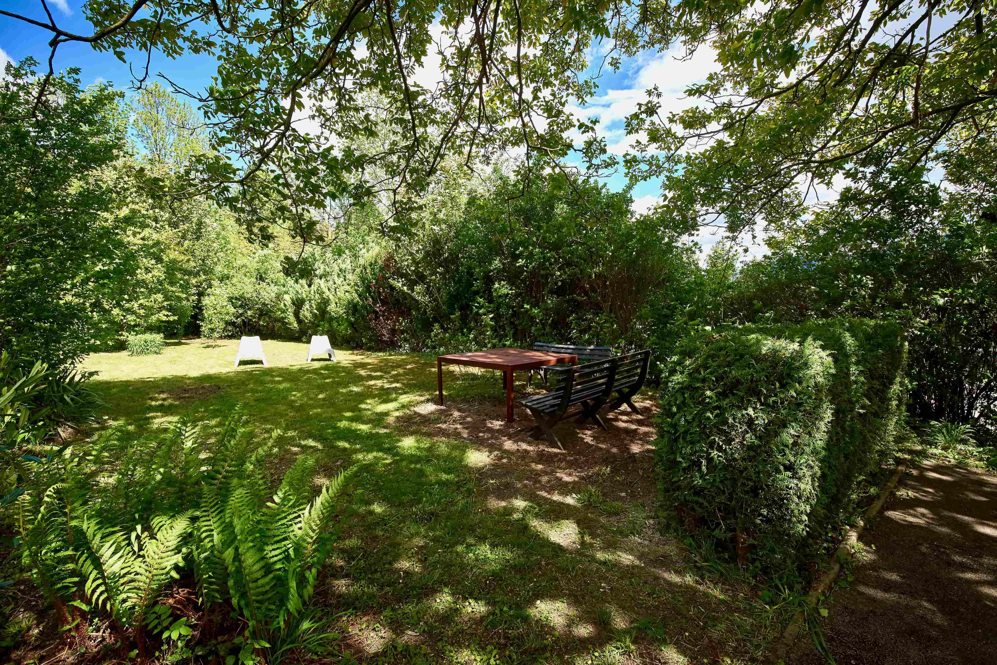 A green garden with trees, shrubs, two white sun loungers and a seating area with a table in the shade.