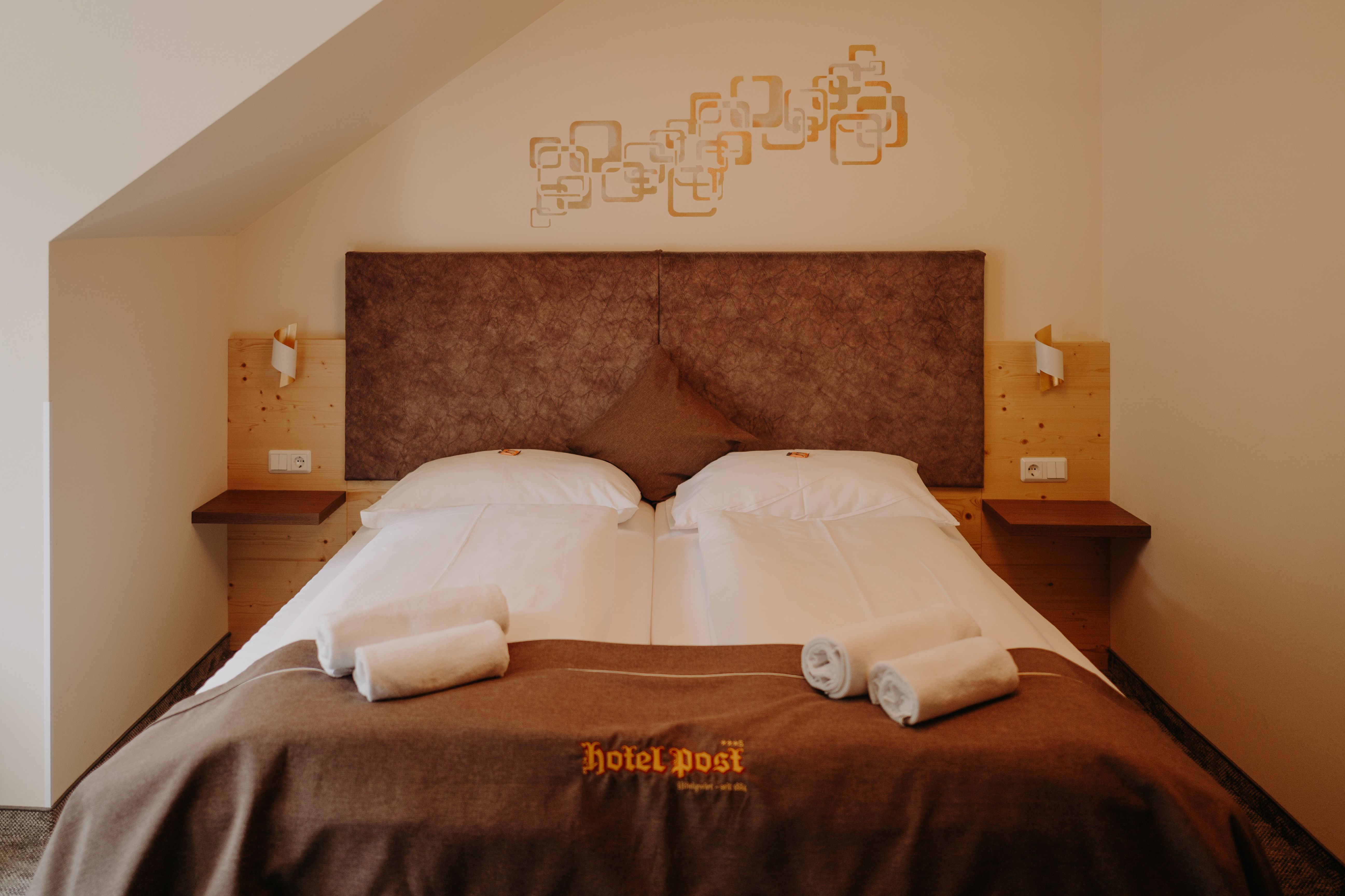 A cozy hotel room with a double bed, featuring light wood paneling and earthy tones.