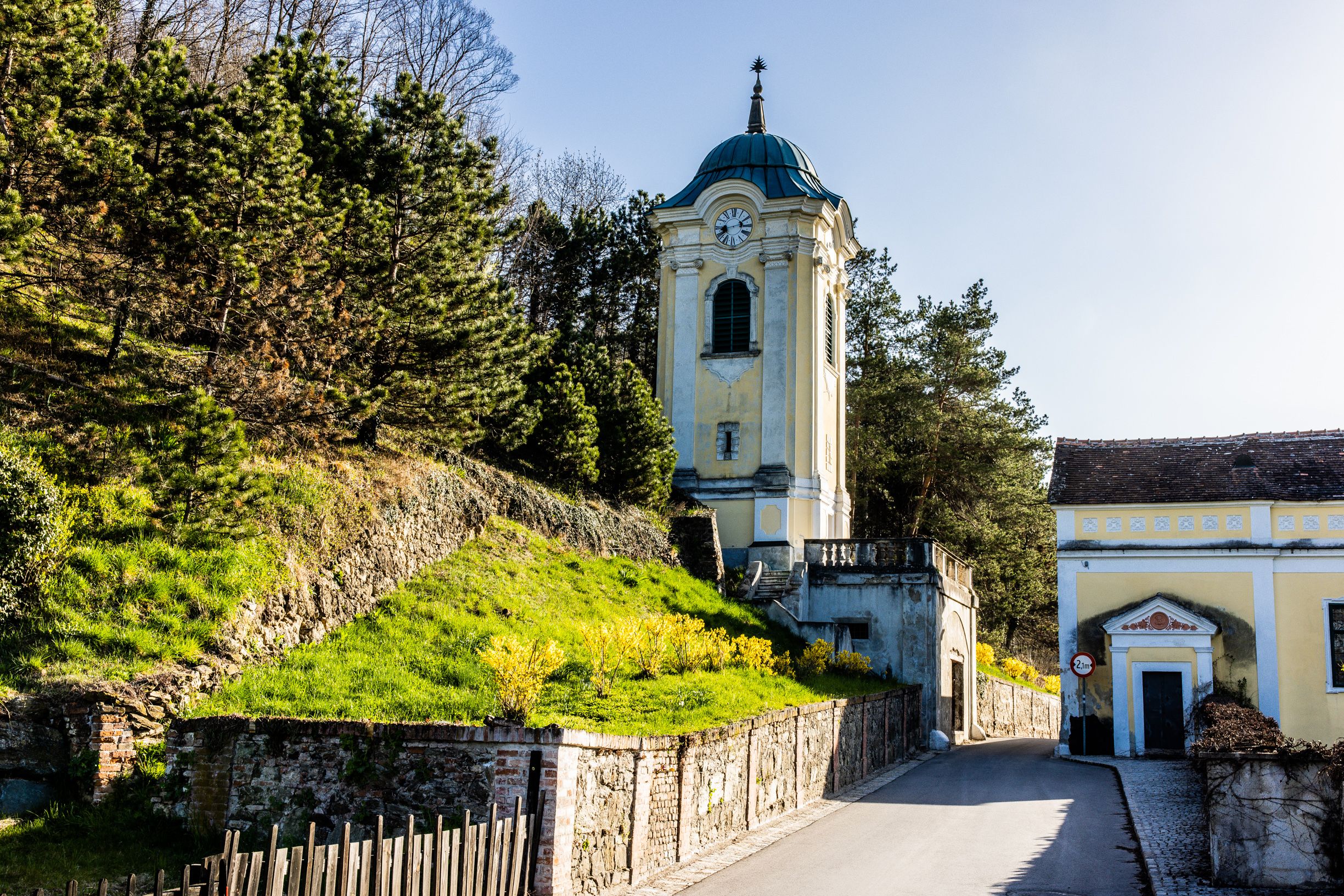 A yellow clock tower in Bad Erlach, surrounded by trees and a sunny slope, a narrow road leads past.