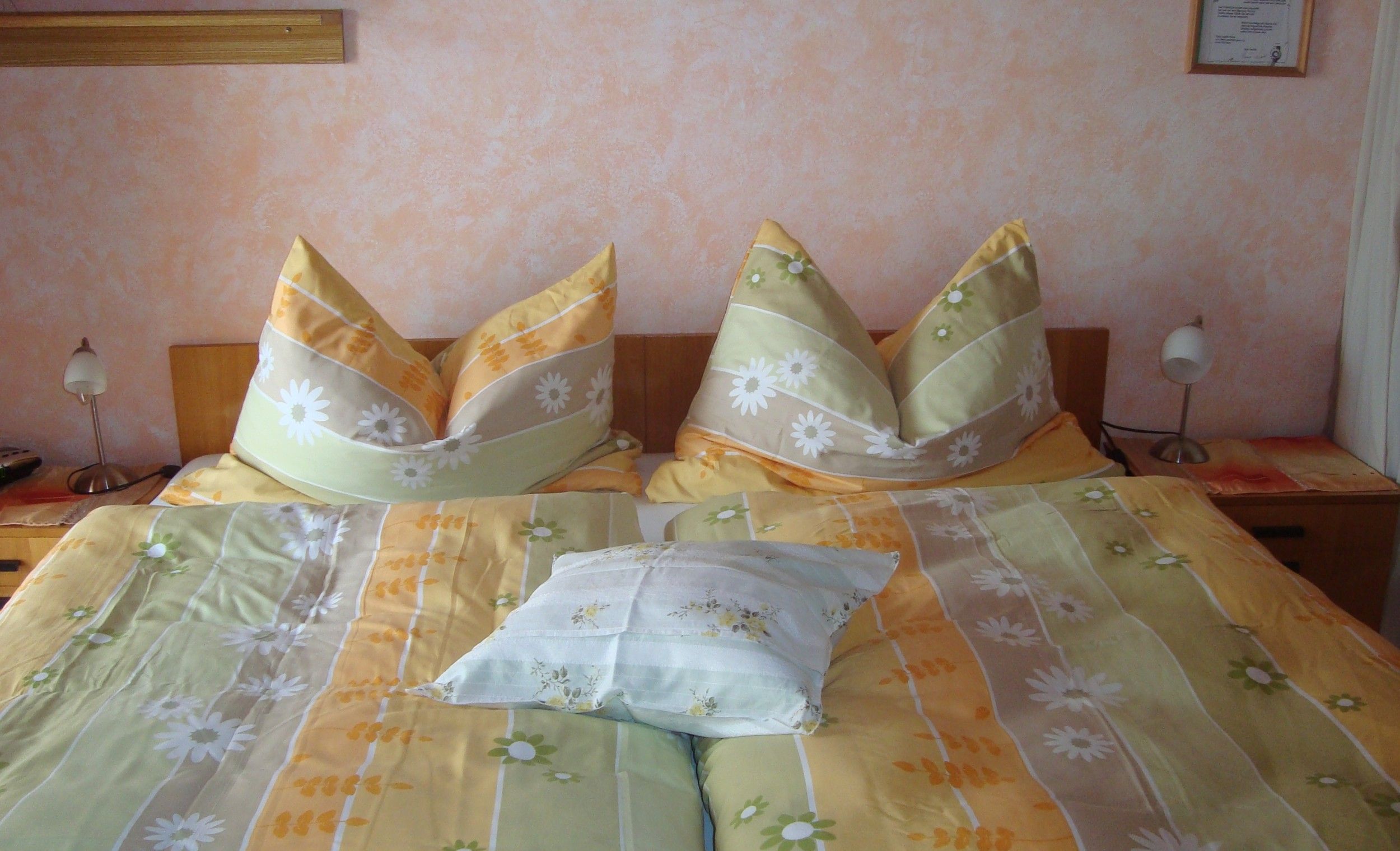 A cozy bedroom with a double bed covered with yellow-green bed linen and floral decor. Two bedside lamps stand next to it.