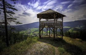 Wooden lookout tower with a view of green valleys and mountains under a blue sky.