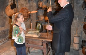 An older man shows a girl a tool in a historical room with old objects.