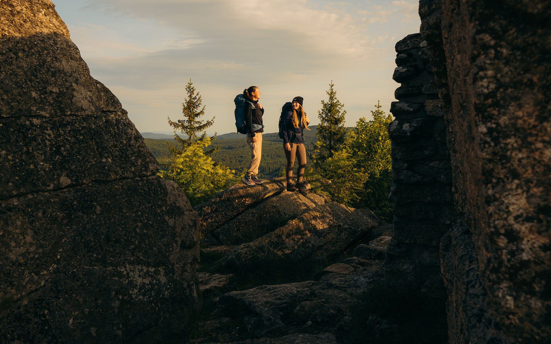 Two hikers stand between imposing rock formations and enjoy the sweeping view of the forest landscape in the golden morning light in the Waldviertel.
