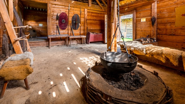 Interior view of a reconstructed Celtic house with wooden walls, a large cauldron over a fireplace and shields on the wall.