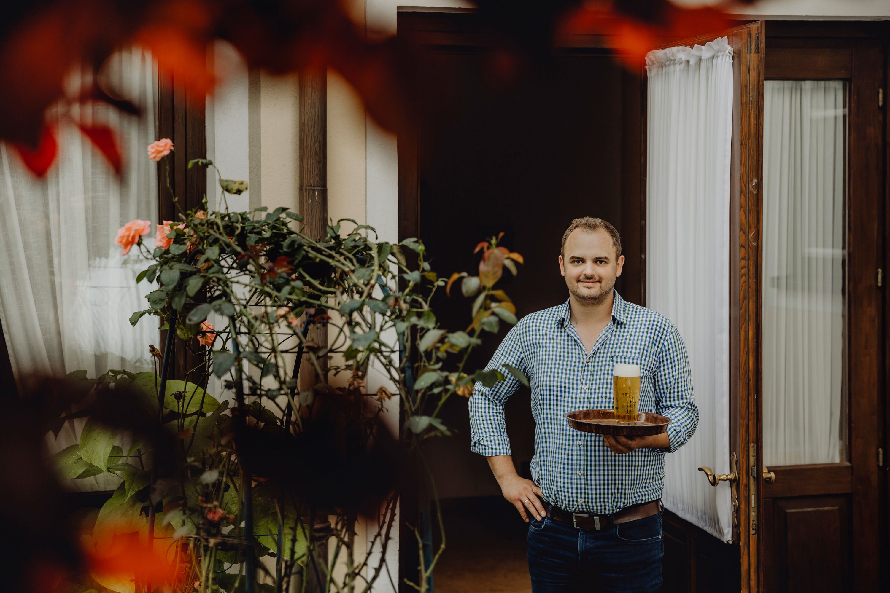 Man with tray and beer in front of a door, surrounded by plants.