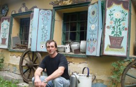Man sitting in front of an old building with painted shutters and old household objects.