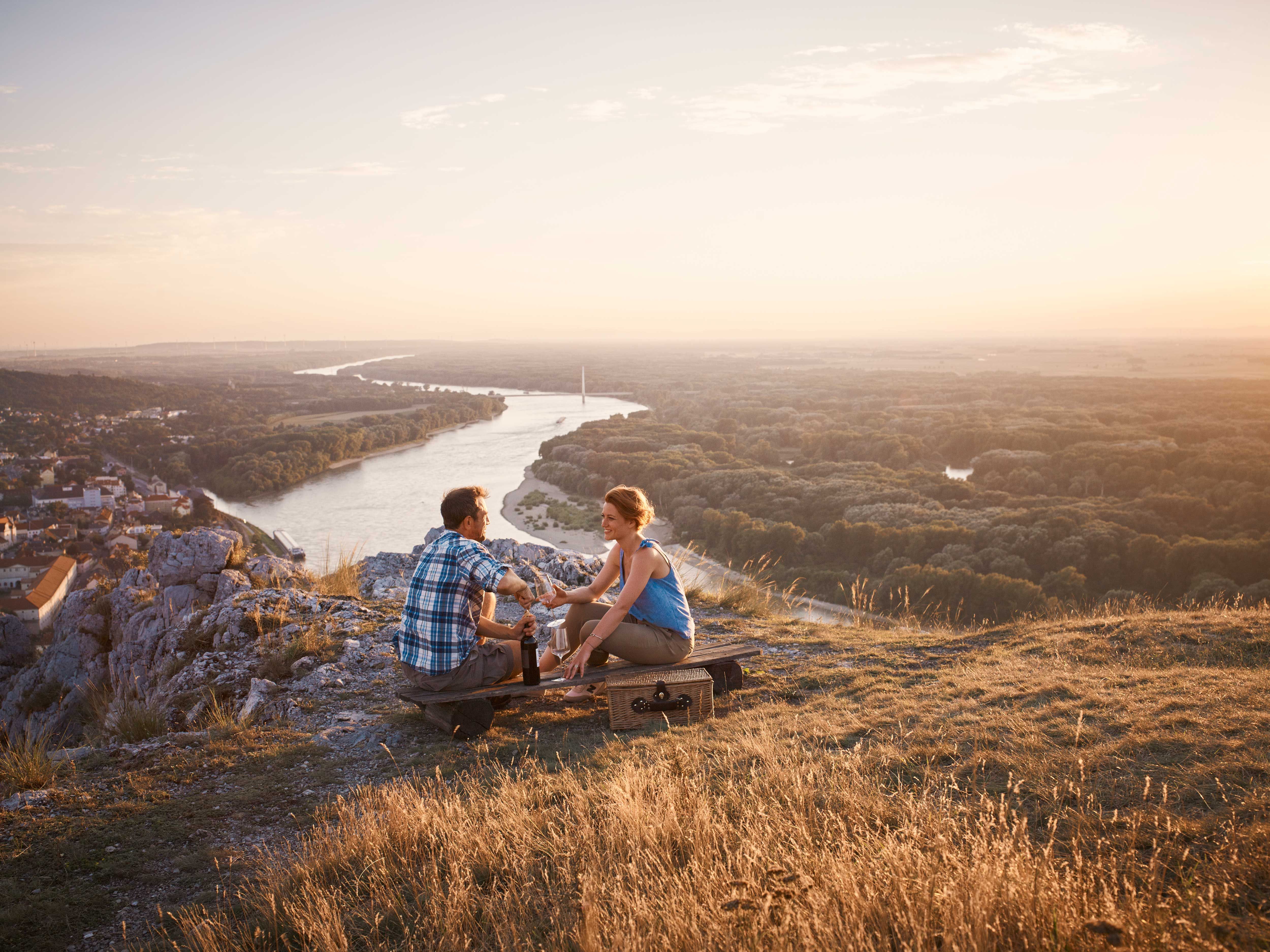 Two people sitting on a hill overlooking a river and a city at sunset.