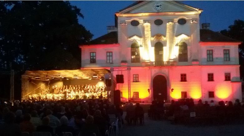 An illuminated building with an orchestra playing on an outdoor stage, surrounded by a seated audience at night.