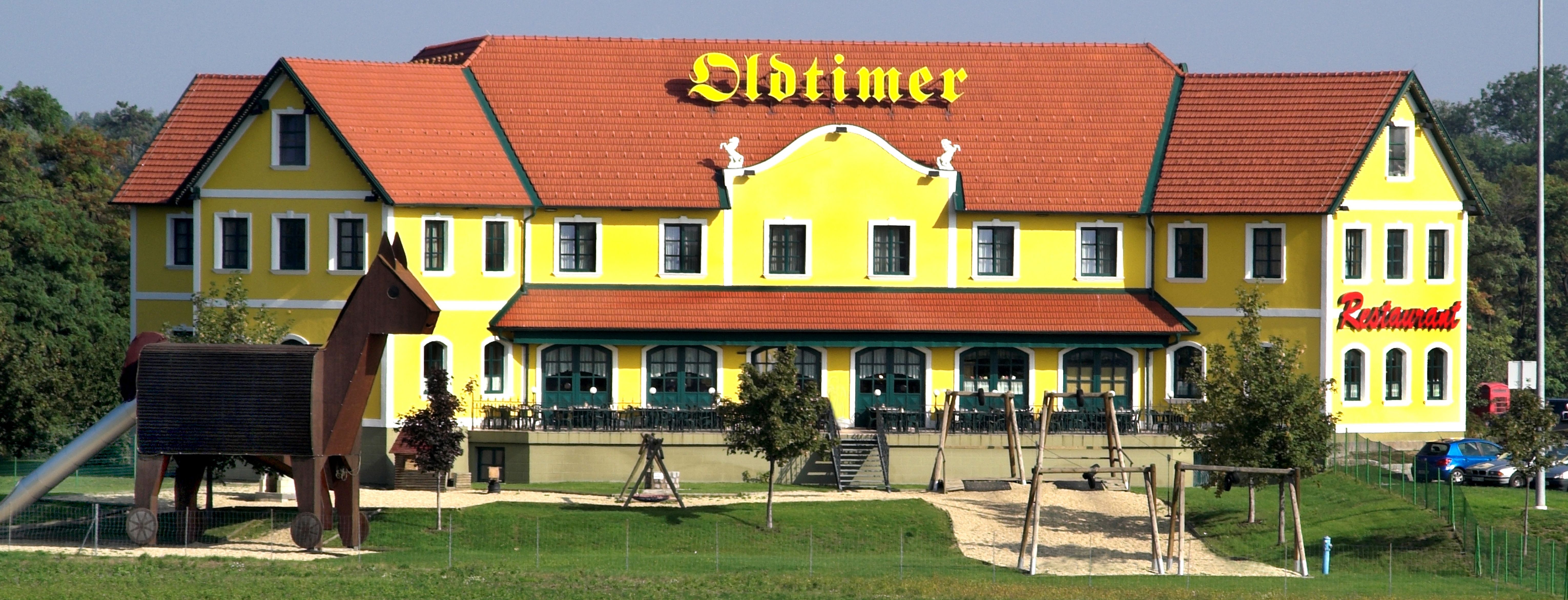 Yellow building with a red roof and the inscription 'Oldtimer', next to it a playground with a large wooden figure.