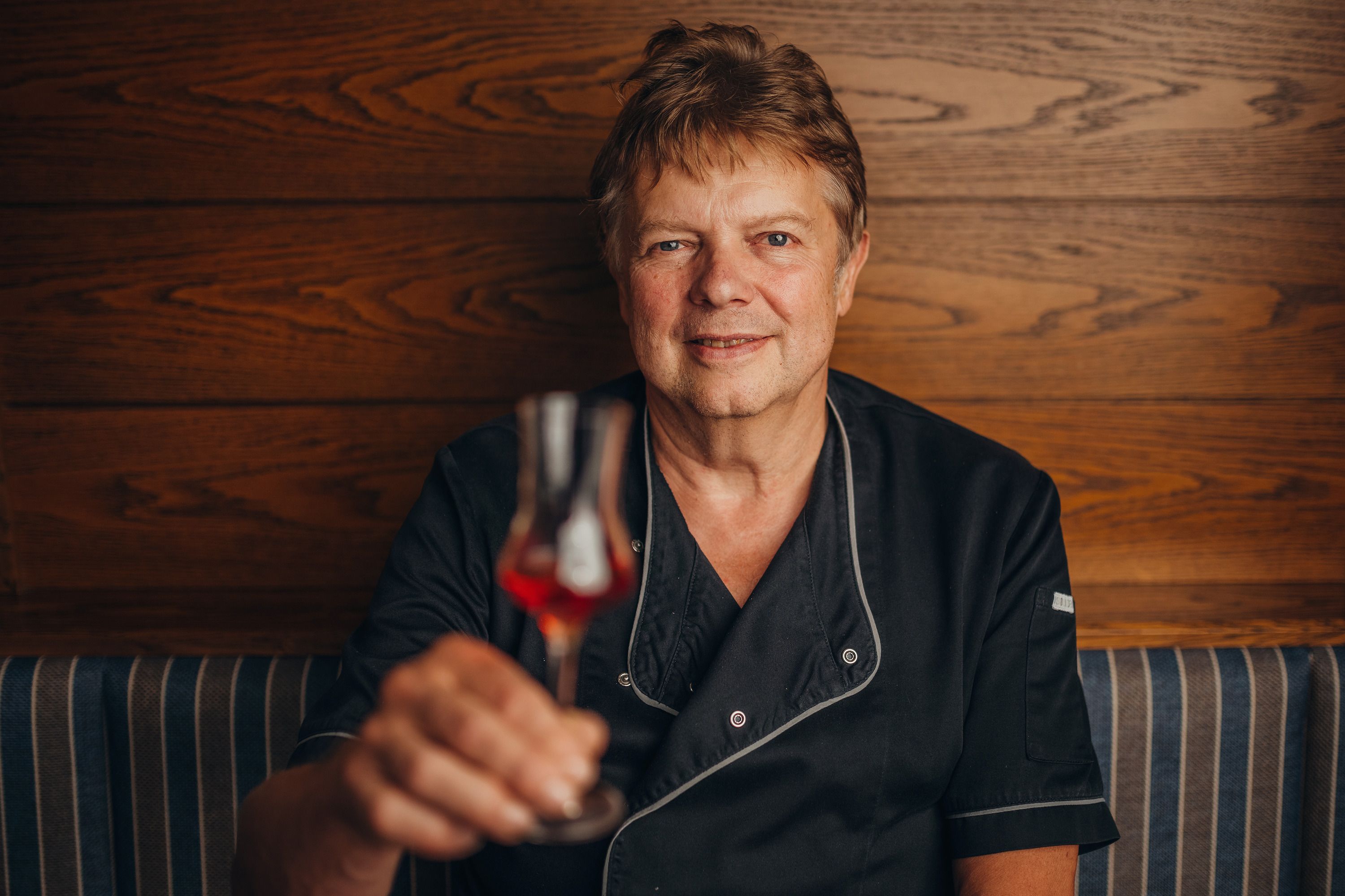 A man in black clothing holds a glass of red drink in front of a wooden wall.
