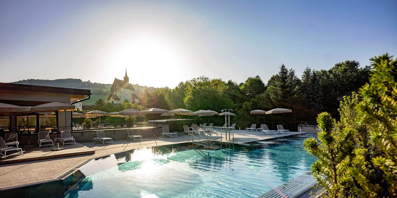 Outdoor area of a hotel with pool, sun loungers and umbrellas, surrounded by trees. 