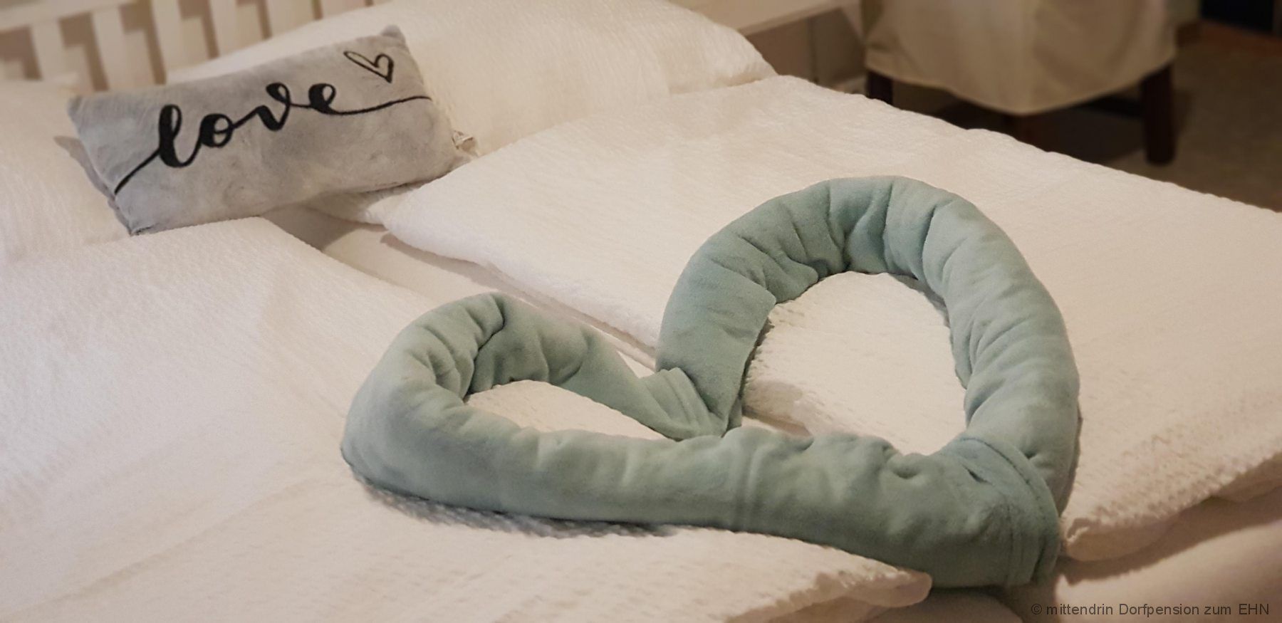 A bed with white pillows and blankets, a gray pillow with the word 'love' on it and a heart-shaped, green fabric pillow.
