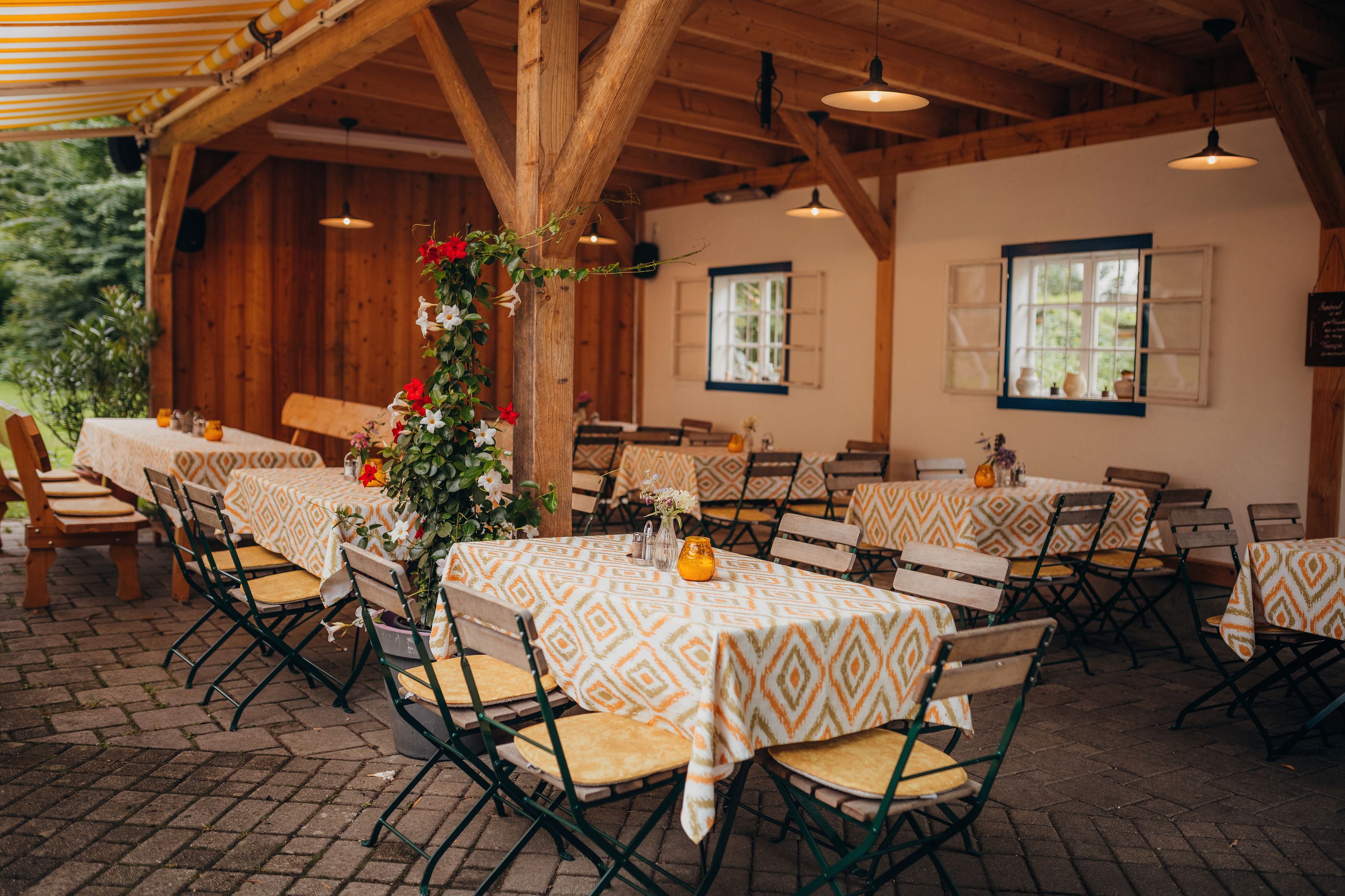A covered guest garden with wooden tables and chairs, decorated with colorful tablecloths and flowers.