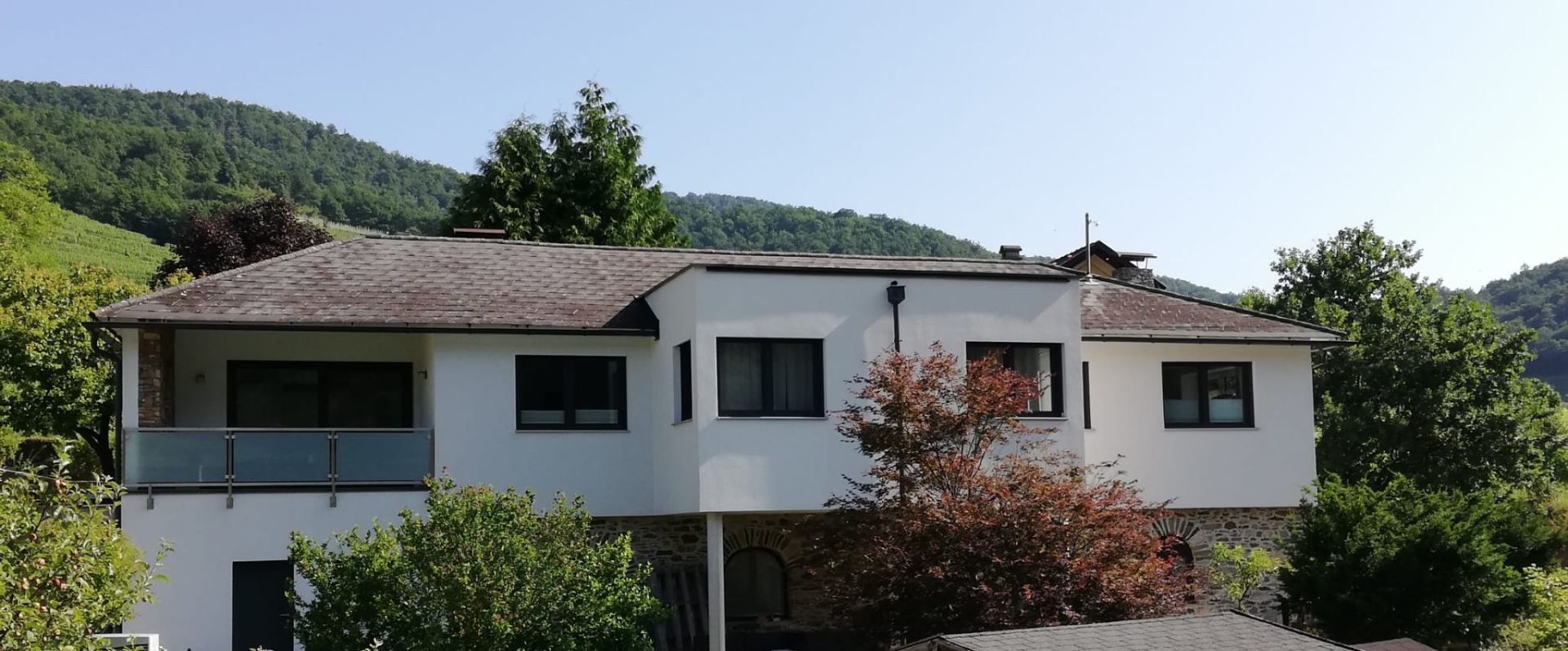 Exterior view of a modern white vacation home with trees in the foreground and wooded hills in the background.