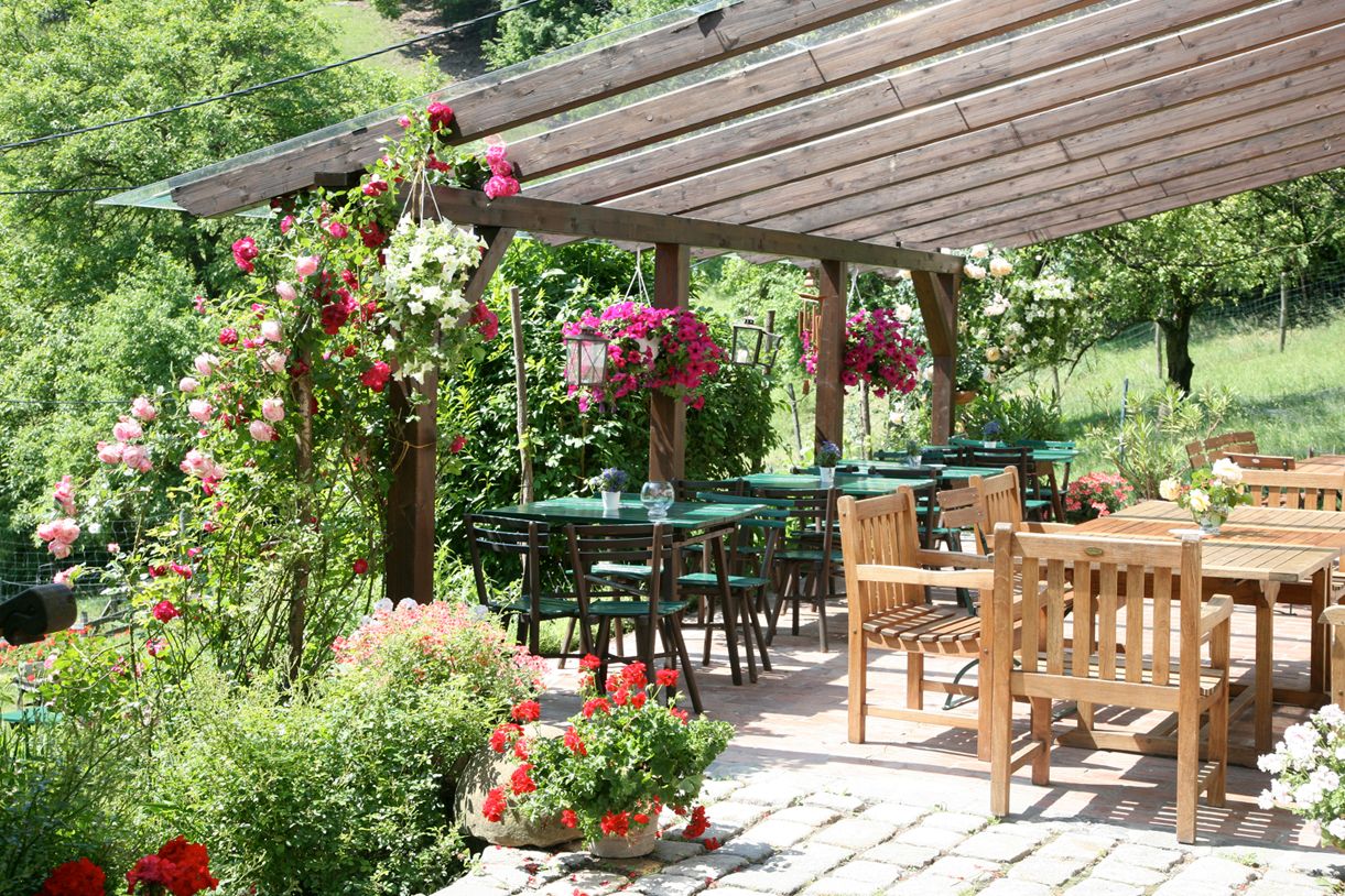 A cozy outdoor area of a wine tavern with wooden tables and chairs, surrounded by flowering plants and a wooden pergola roof.