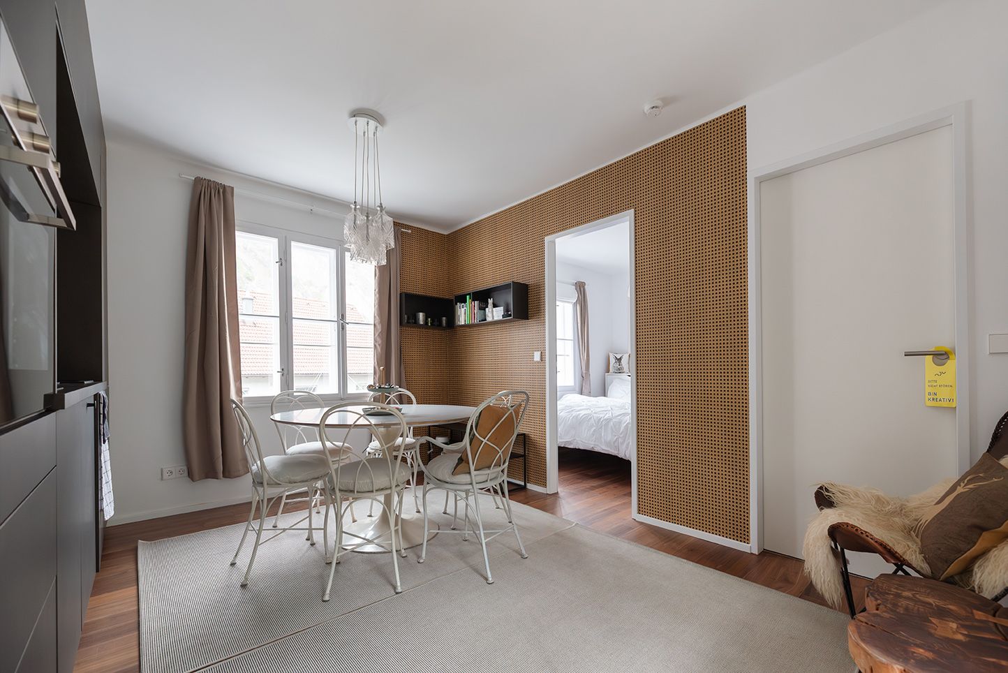 Modern furnished living room with dining table and chairs, wooden wall and view into the bedroom.
