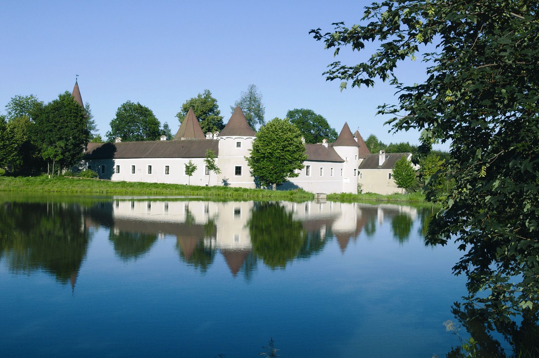 The castle with its pointed towers is reflected in a calm lake surrounded by trees.