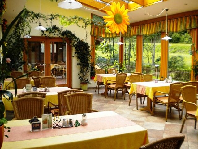 A cozy winter garden with yellow tablecloths, rattan furniture and plants. Large windows offer a view of the greenery. A large sunflower hangs from the ceiling.