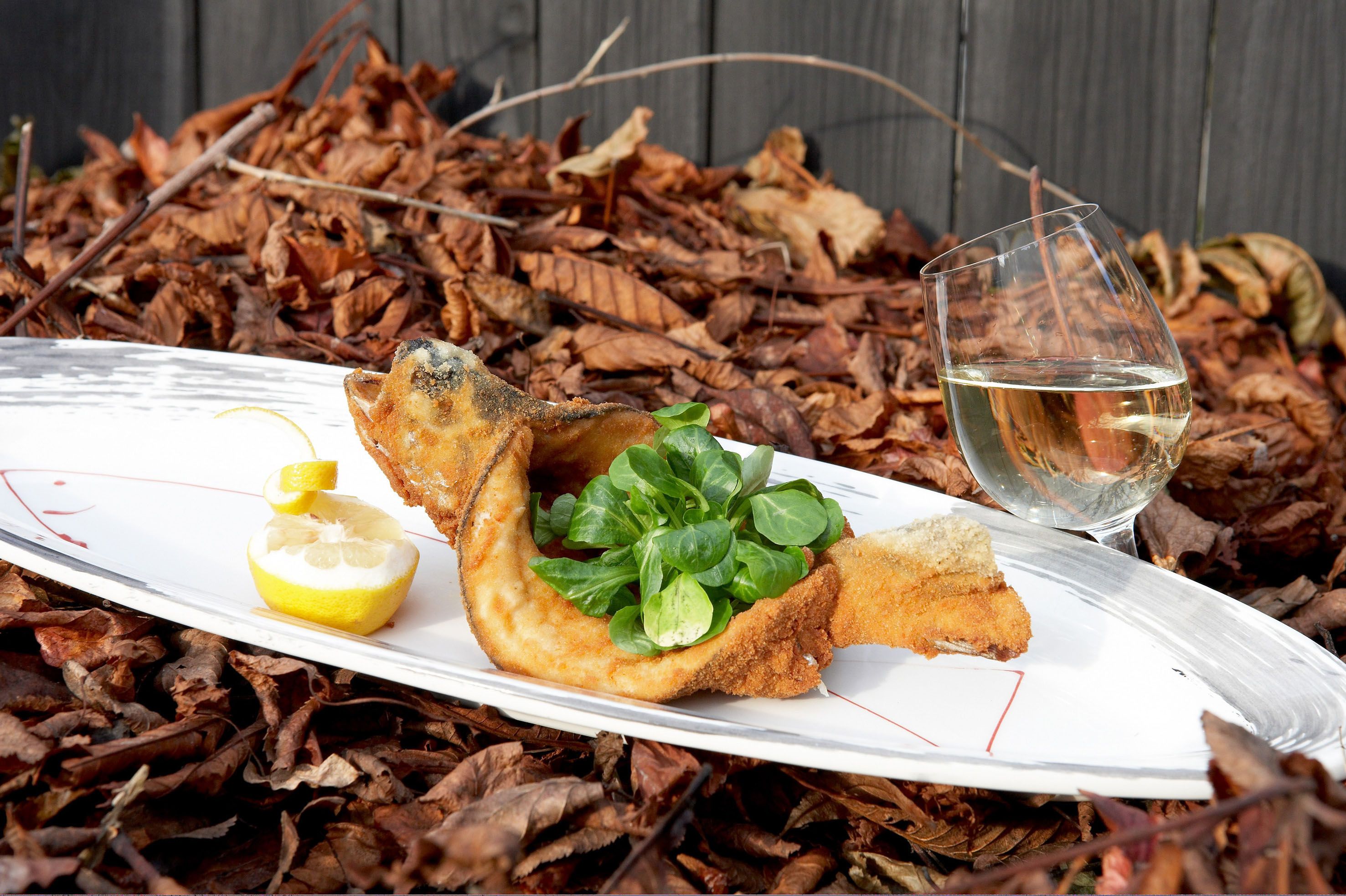 Fried fish with lamb's lettuce and lemon on a plate, next to a glass of white wine, on a bed of autumn leaves.