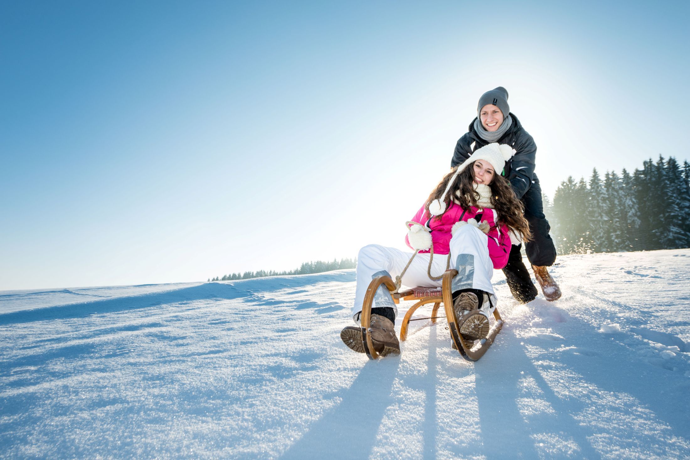 Two people are happily tobogganing down a snowy hill under a clear sky.