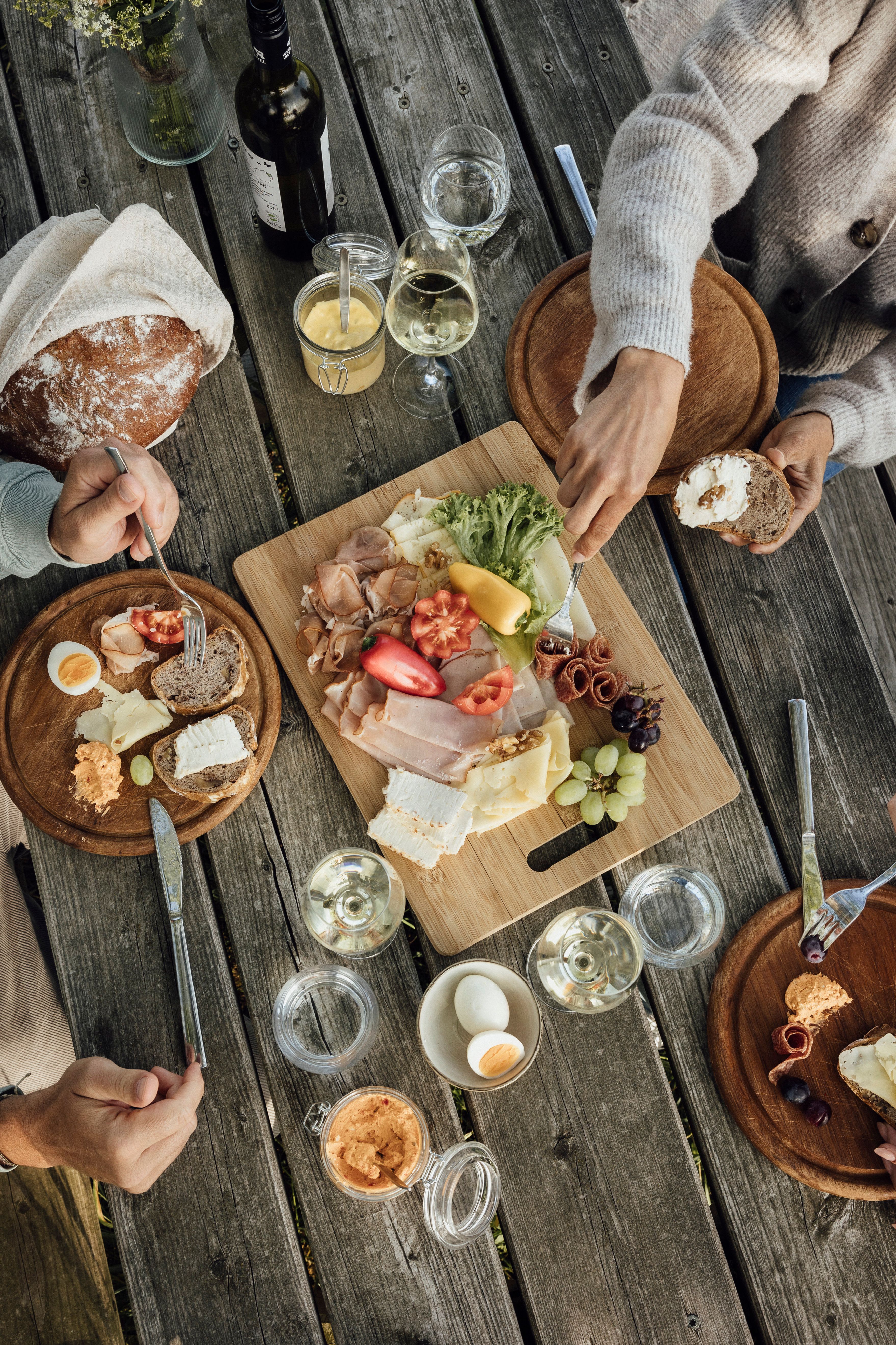 A rustic wooden table with a snack of bread, cheese, cold cuts, vegetables and wine.