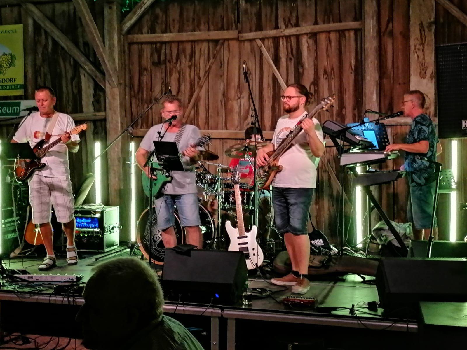 A band plays on a stage in a rustic wooden building. Five musicians with guitars, drums and keyboards can be seen.