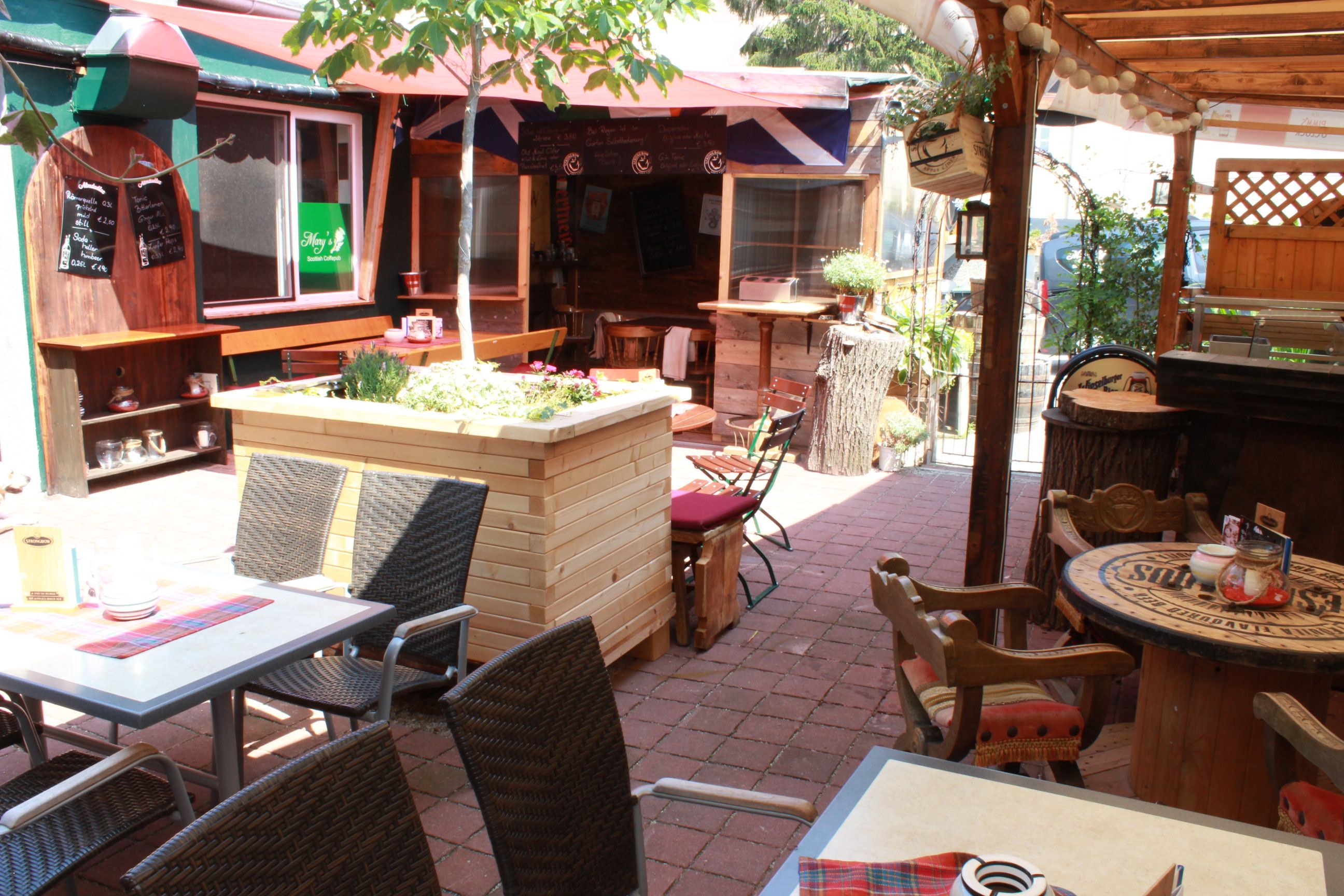 A cozy pub garden with tables, chairs and plants under a sun canopy.
