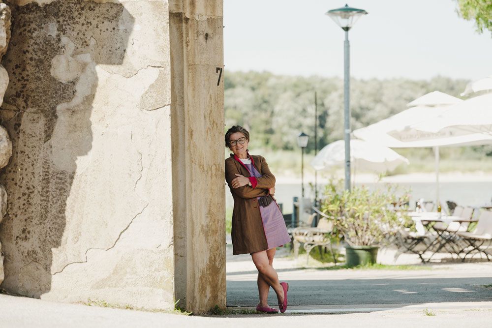 Smiling woman leaning against a wall, in the background a terrace with tables, chairs and parasols.