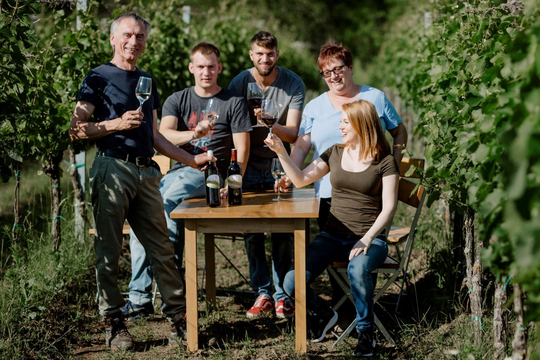 A group of five people sit and stand at a table in the vineyard, holding wine glasses and smiling.