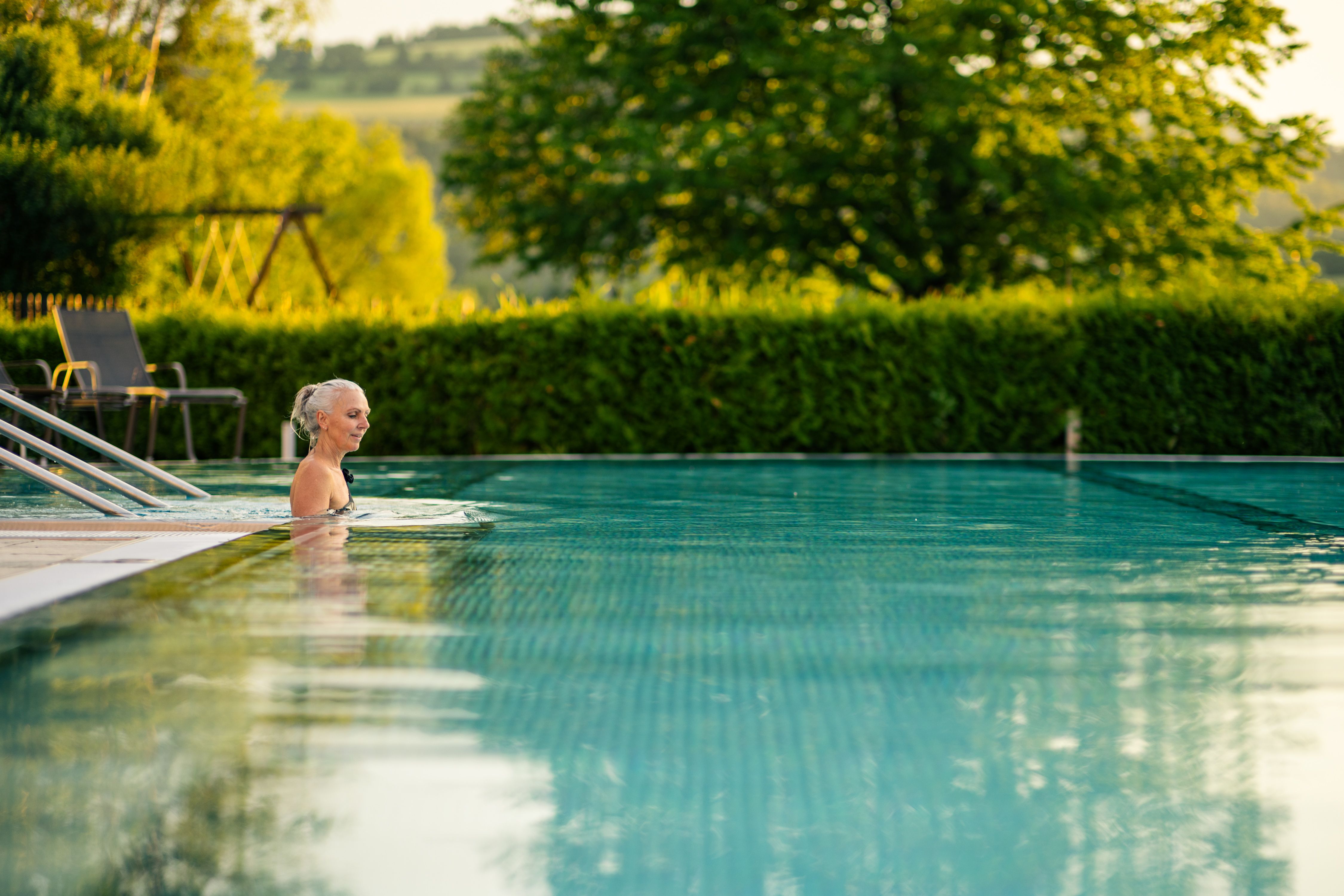 A woman is getting into the outdoor pool with a green landscape in the background.