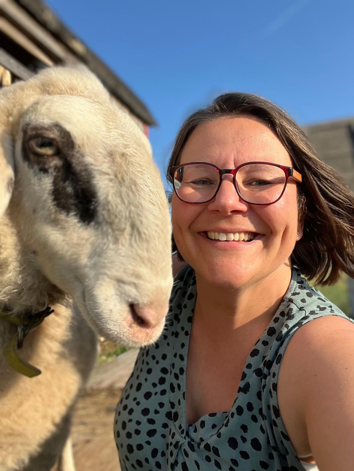 Woman with glasses smiles next to a sheep in the sun.