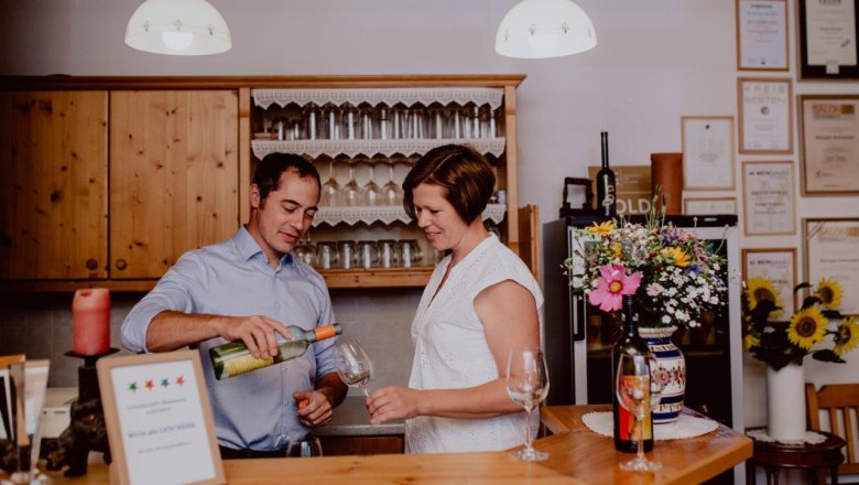 A man pours wine for a woman at a bar. Glasses and awards can be seen in the background.