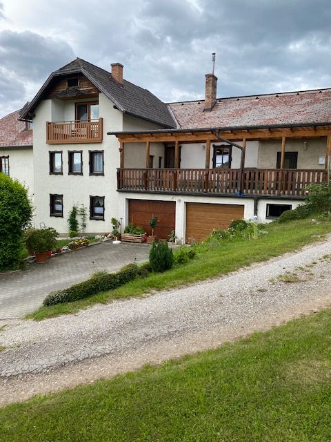 A two-storey house with a balcony and garage, surrounded by green countryside and a cloudy sky.