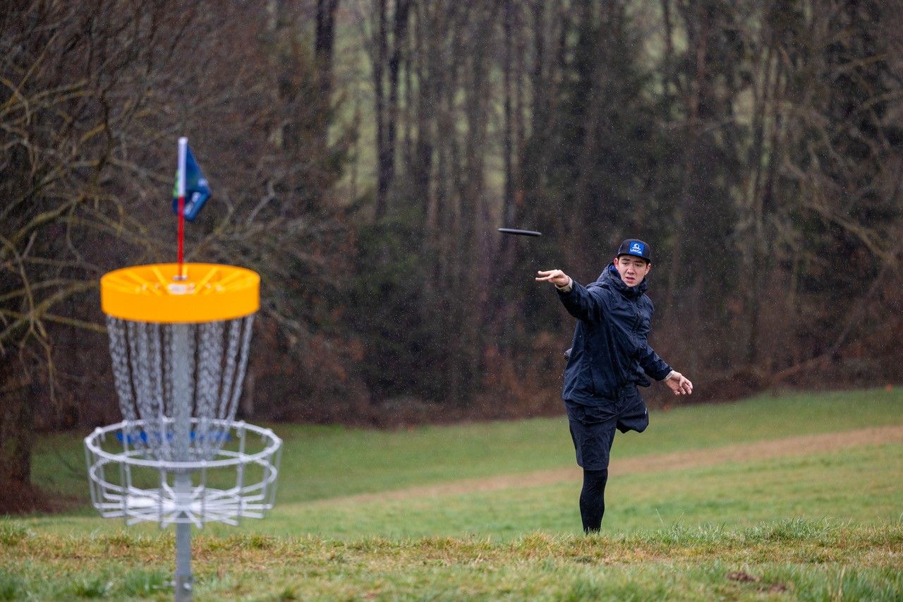 A man throws a disc towards a disc golf basket in a meadow surrounded by trees.