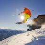A skier jumps into the air in front of the Terzerhaus on the Gemeindealpe, surrounded by snow-covered mountains and a blue sky.