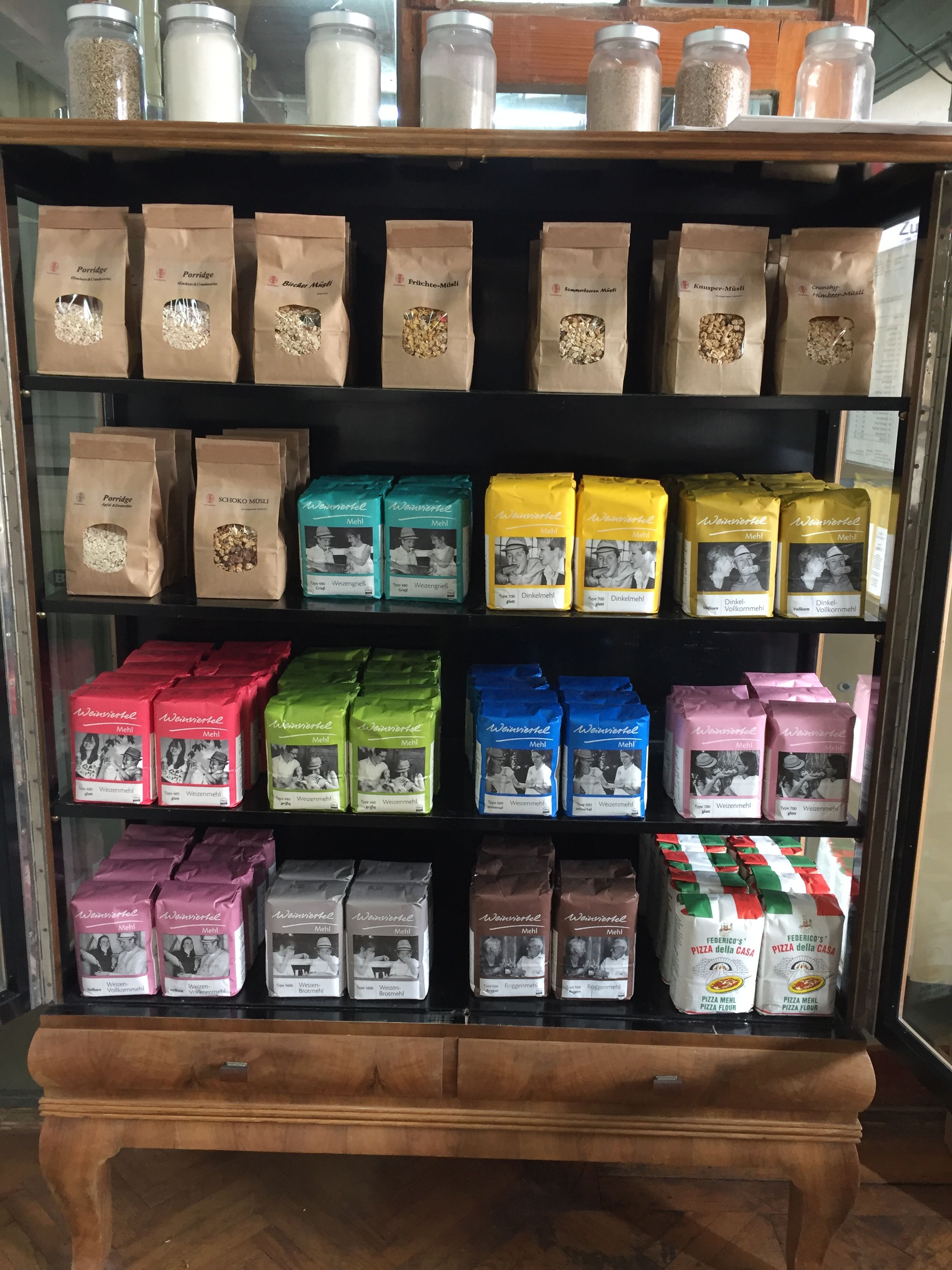 Shelf with different types of flour in colorful packaging and jars of cereal on top.