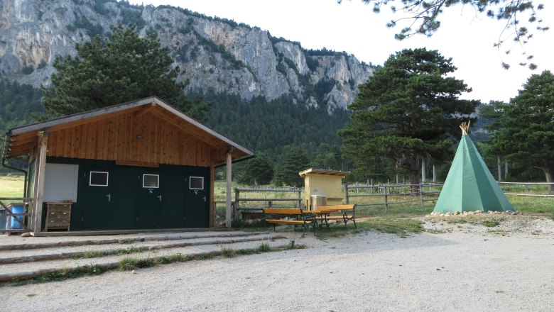 A campsite with a green tepee, a wooden hut and benches in front of a rock face and trees.