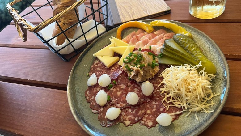 A plate with cold cuts, cheese, gherkins and onions, next to it a basket of bread and a drink on a wooden table.
